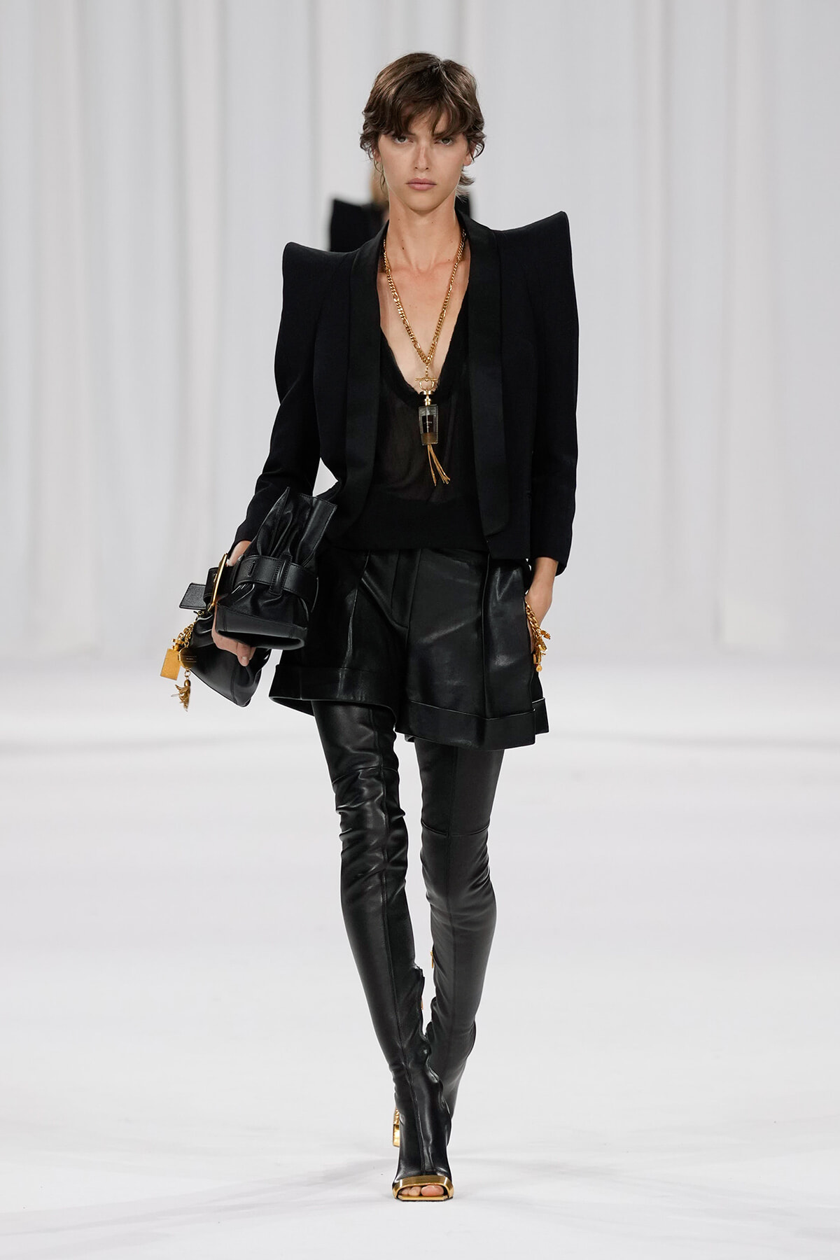 Fashion model on a runway in an all-black, high-fashion outfit with a structured blazer, leather shorts, and thigh-high boots, accessorized with gold jewelry and a bag.