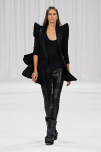 Model walking a white runway in an all-black outfit: structured blazer with exaggerated shoulders, black top, leather-paneled pants, and tall black boots.