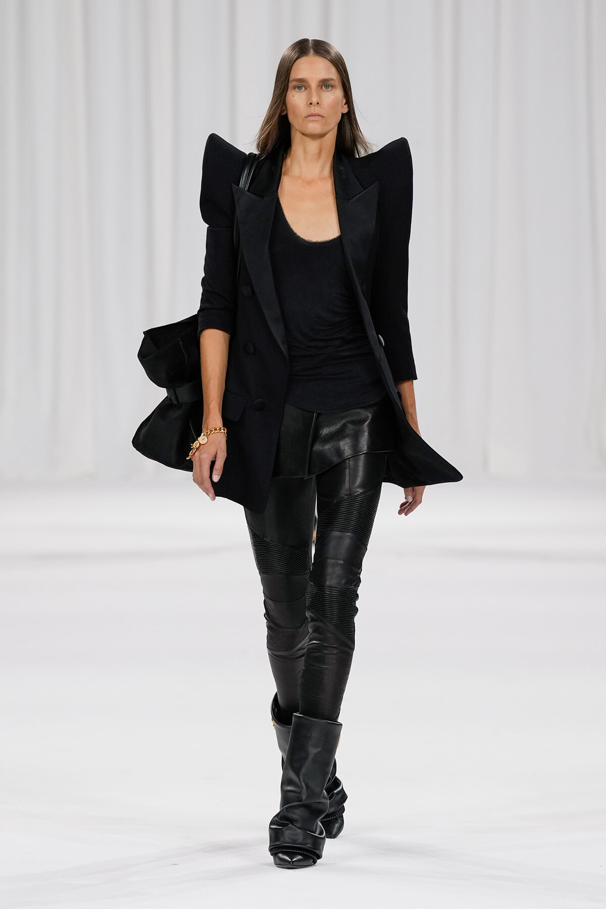 Model walking a white runway in an all-black outfit: structured blazer with exaggerated shoulders, black top, leather-paneled pants, and tall black boots.