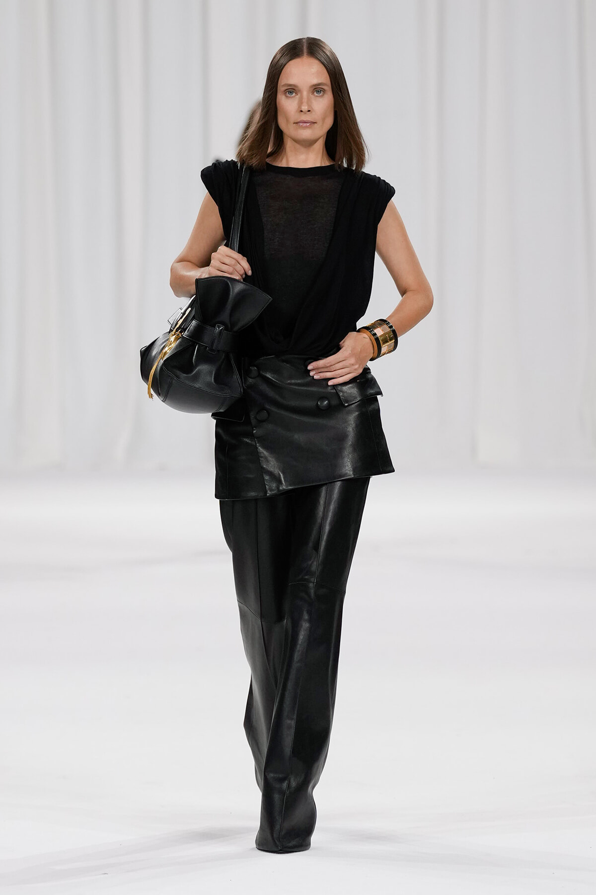 Model walking the runway in an all-black leather ensemble: draped top, layered skirt over wide-leg leather pants, and a black handbag.
