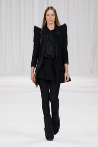 Model on a runway wearing an all-black, structured blazer with exaggerated shoulders over a black blouse and slim pants with stacked black boots.
