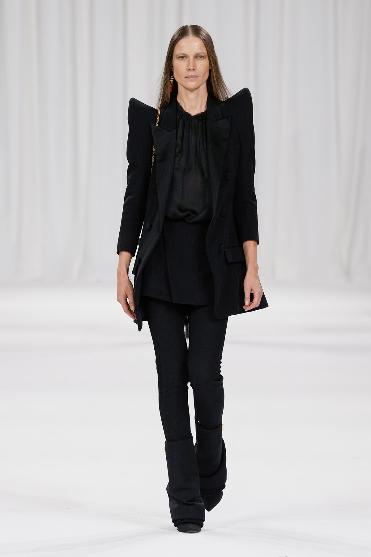 Model on a runway wearing an all-black, structured blazer with exaggerated shoulders over a black blouse and slim pants with stacked black boots.
