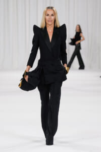 Runway model walks in a black tailored pantsuit with dramatic padded shoulders, holding a black bag with gold hardware.