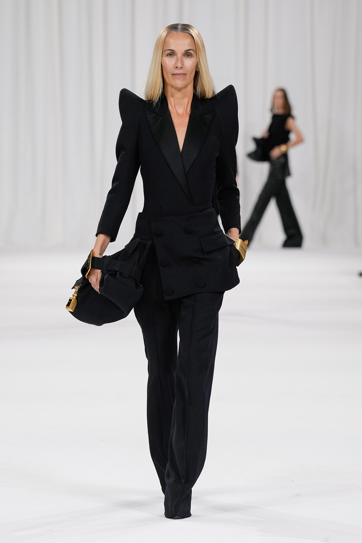 Runway model walks in a black tailored pantsuit with dramatic padded shoulders, holding a black bag with gold hardware.