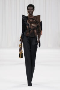 Runway model in a black pantsuit with a sculptural brown sequined top, carrying a small gold handbag and wearing a chunky gold bracelet.
