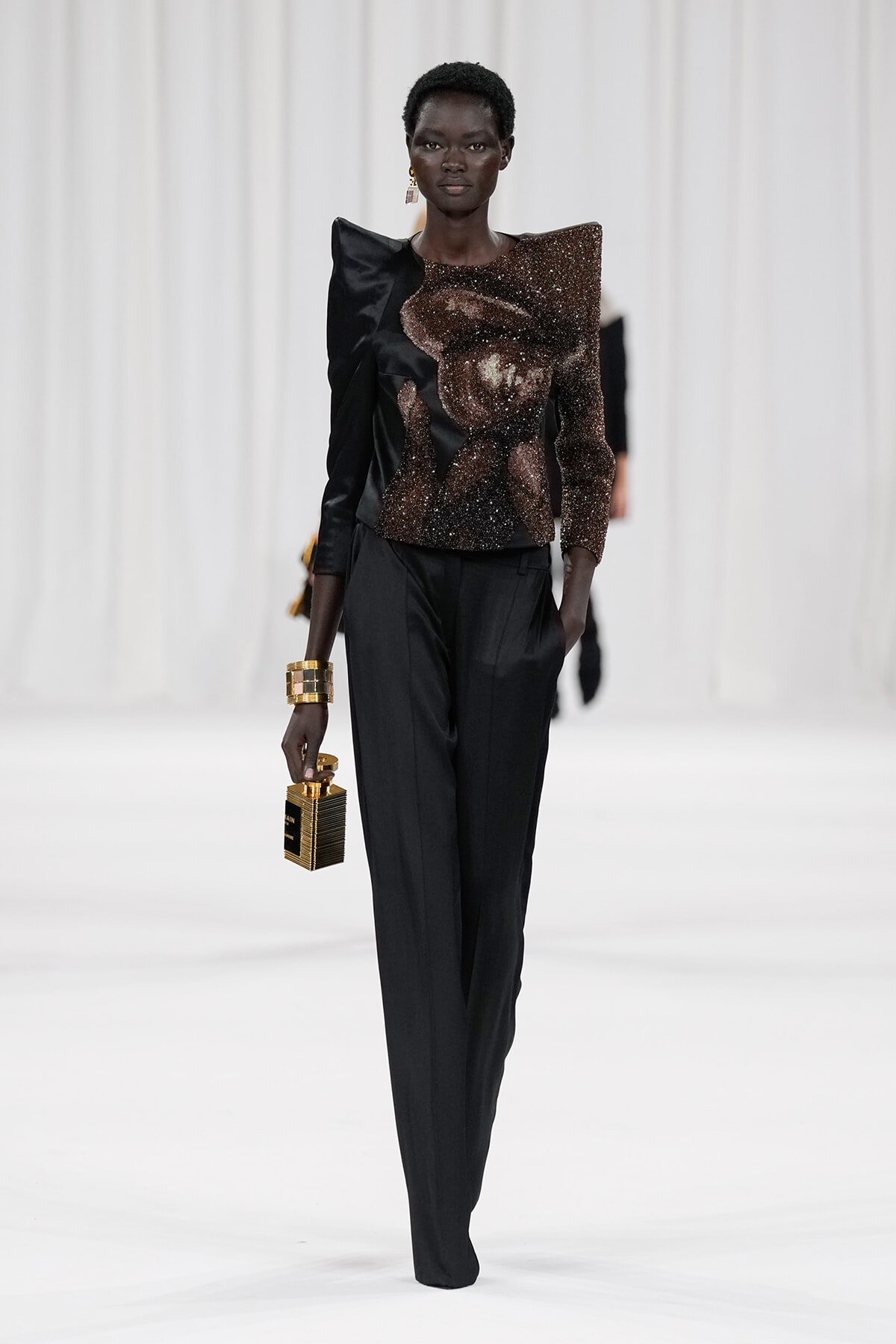 Runway model in a black pantsuit with a sculptural brown sequined top, carrying a small gold handbag and wearing a chunky gold bracelet.