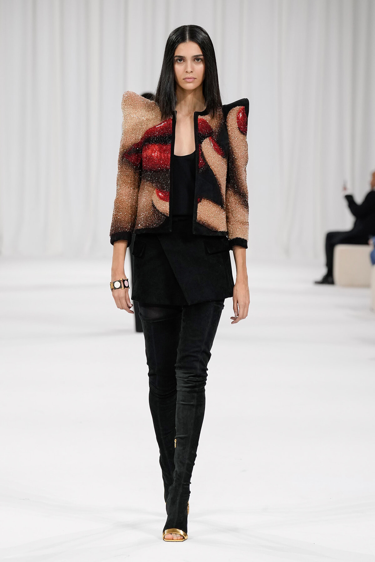 Model on a white runway wearing a glittery, color-block jacket over a black top and black pants with tall boots.