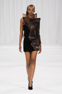 Model walking the runway in a black mini dress with a sparkling brown sheer one-shoulder overlay and black heels.