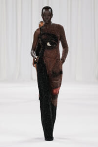 Model on a white runway wearing a two-tone, glittery, asymmetrical gown with black and burgundy sequins.