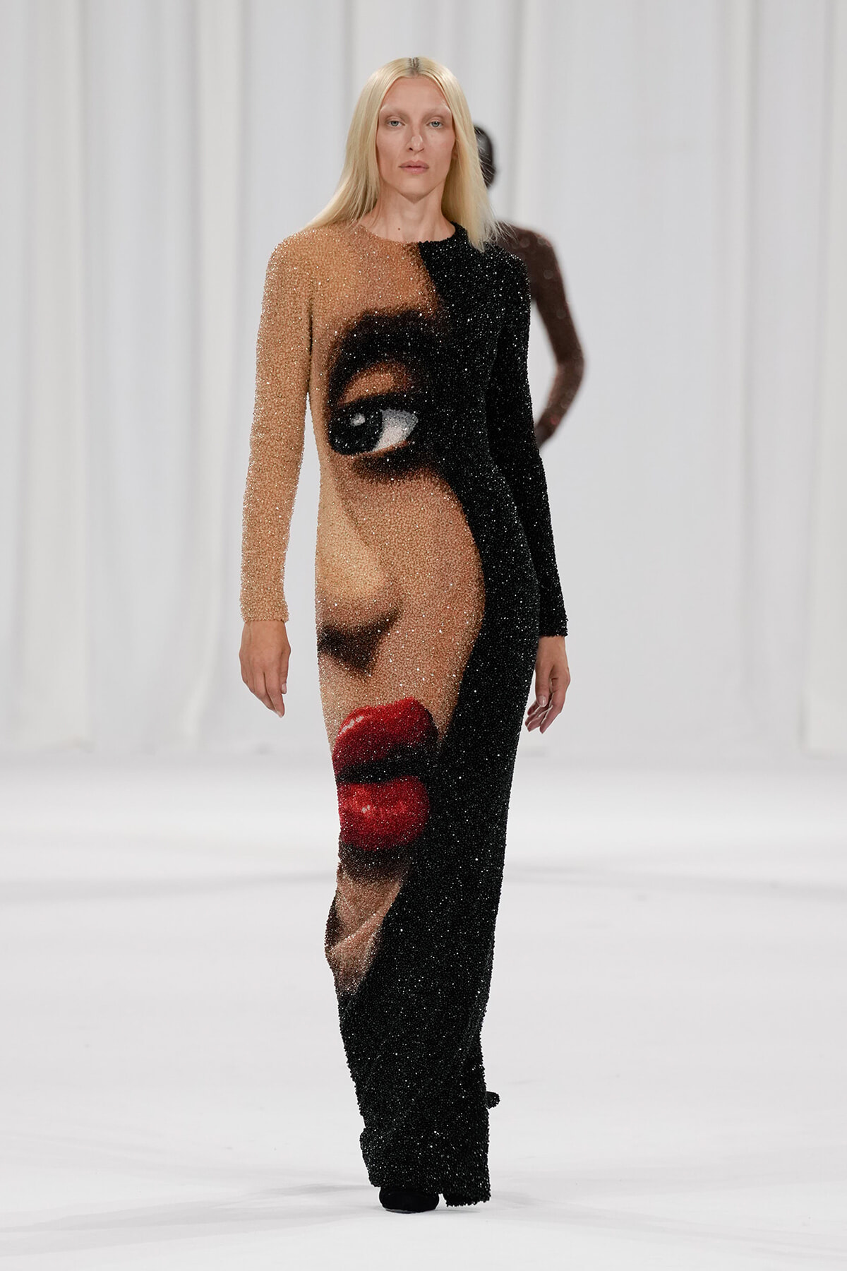 Model walking the runway in a full-length, glittery gown featuring a large portrait of a face across the front (beige and black halves).