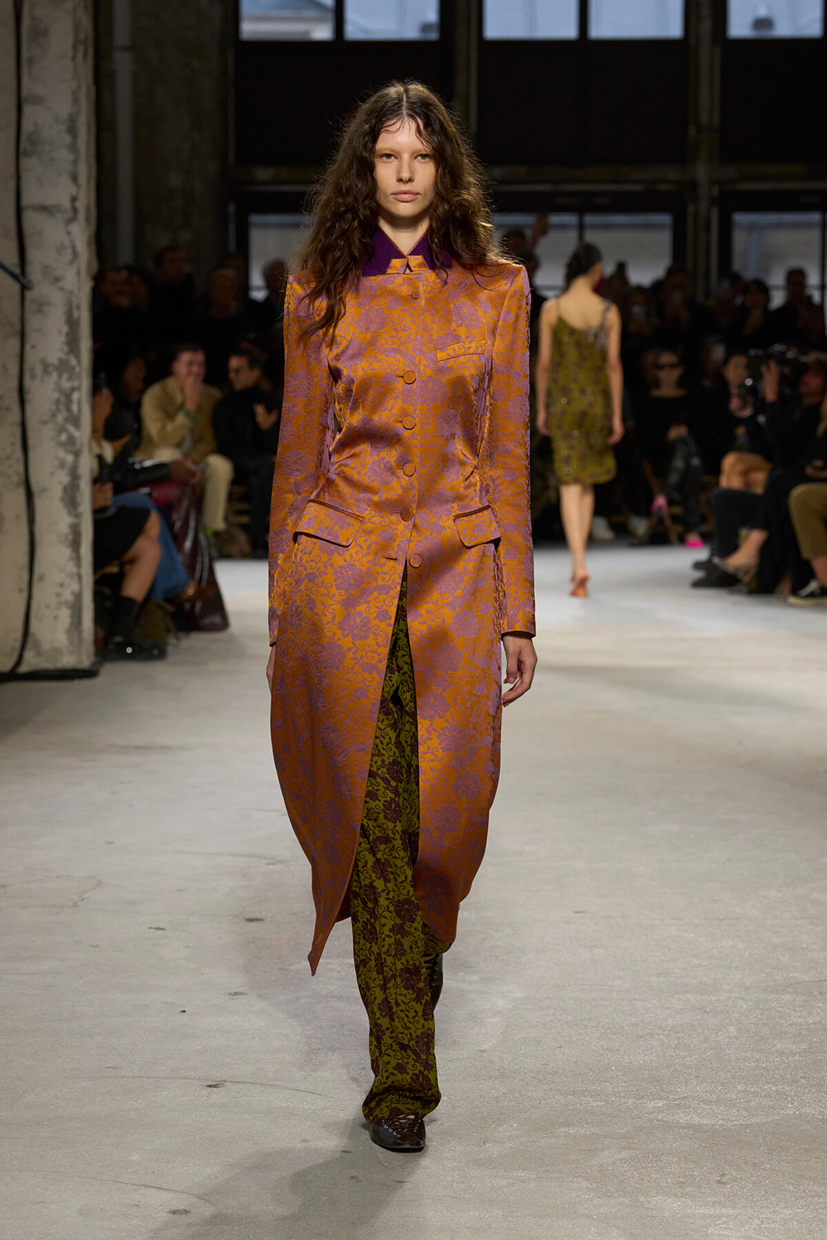 Model walking a fashion runway in a metallic orange-purple brocade suit with a long coat and slit revealing green patterned pants behind.