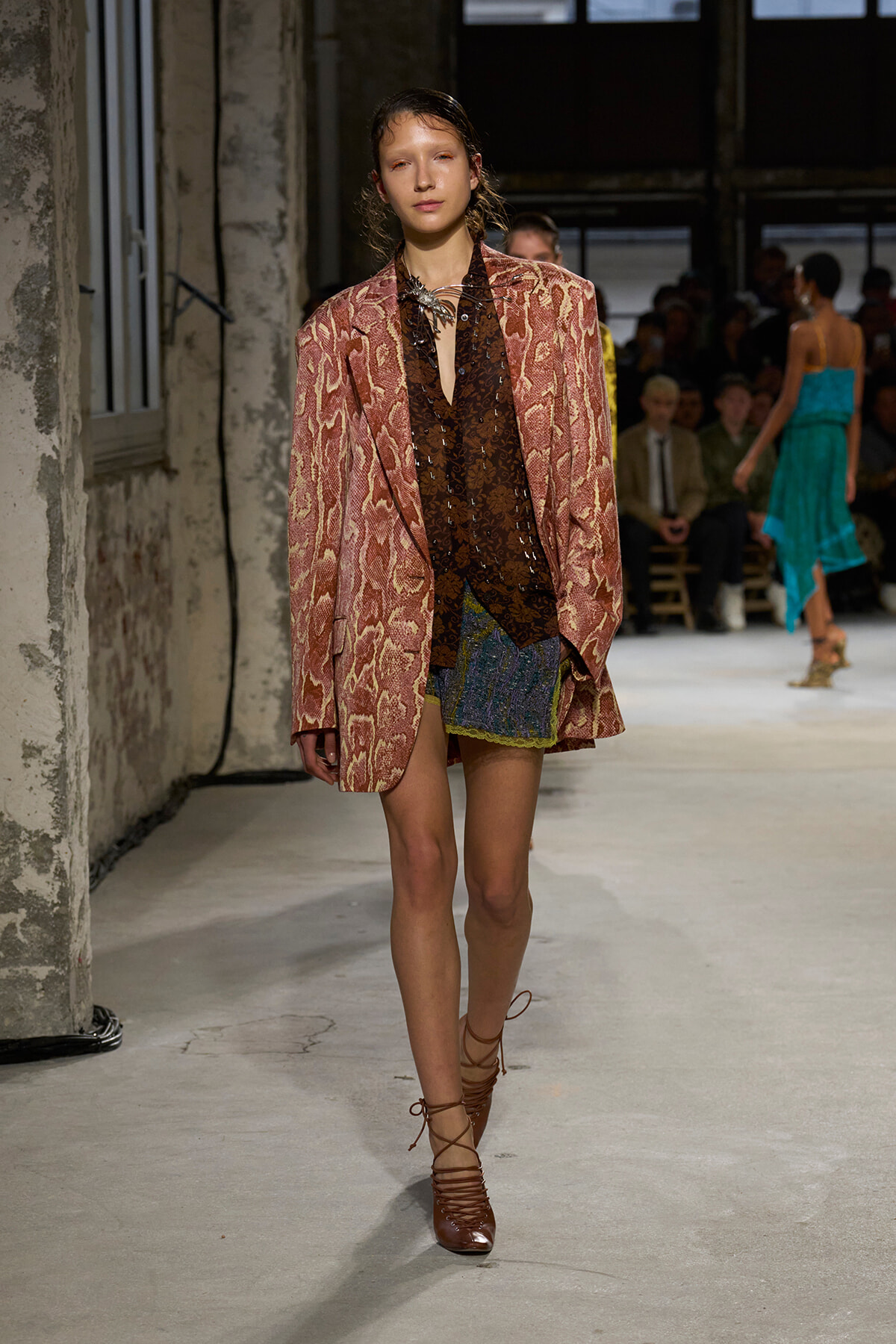 Model on a runway wearing a pink snakeskin blazer over a brown sequined top and a short multicolored skirt; brown lace-up heels.