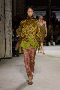 Model walks a fashion runway in a gold floral jacquard blouse with a high collar, paired with green layered hem and pink snakeskin-pattern pants, glittery sandals; audience in the background.