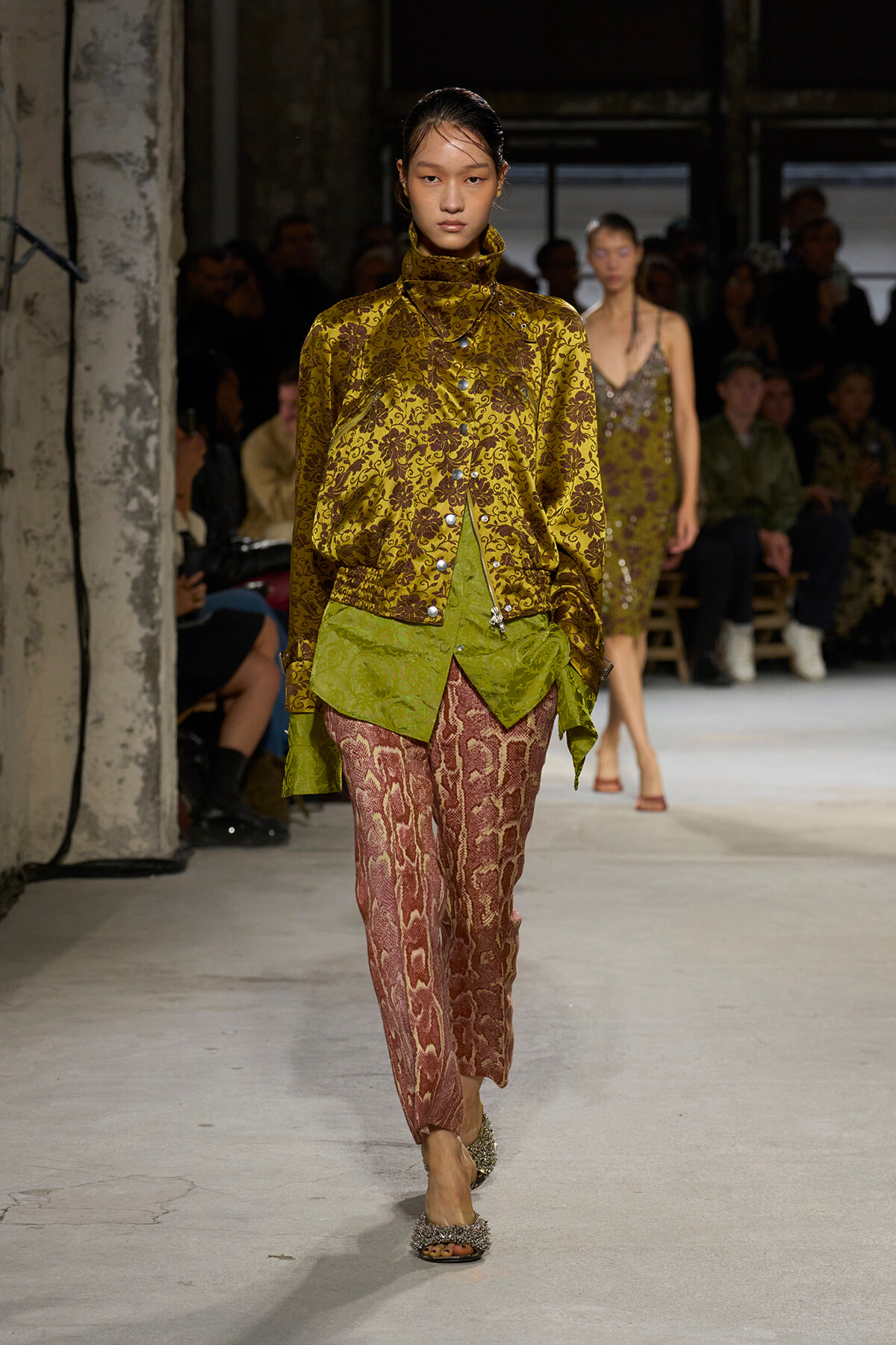 Model walks a fashion runway in a gold floral jacquard blouse with a high collar, paired with green layered hem and pink snakeskin-pattern pants, glittery sandals; audience in the background.