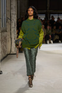 Model walking a fashion runway in an oversized green patterned knit over a lime shirt, metallic green pants, and strappy metallic shoes, carrying a textured bag.