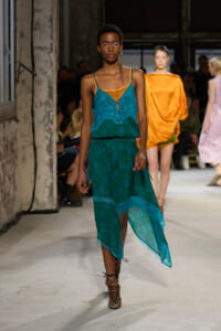 Model on a fashion runway wearing a teal lace dress with orange underlayer and a high-low hem, paired with strappy heels.