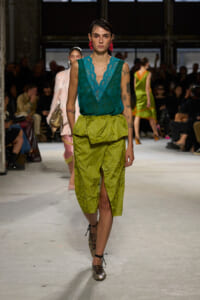 Model on a runway wearing a teal lace V-neck top and lime green brocade skirt with a front slit.