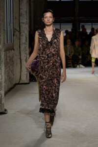 Model walks a fashion runway in a pink-brown lace slip dress, holding a metallic purple bag, in an industrial venue