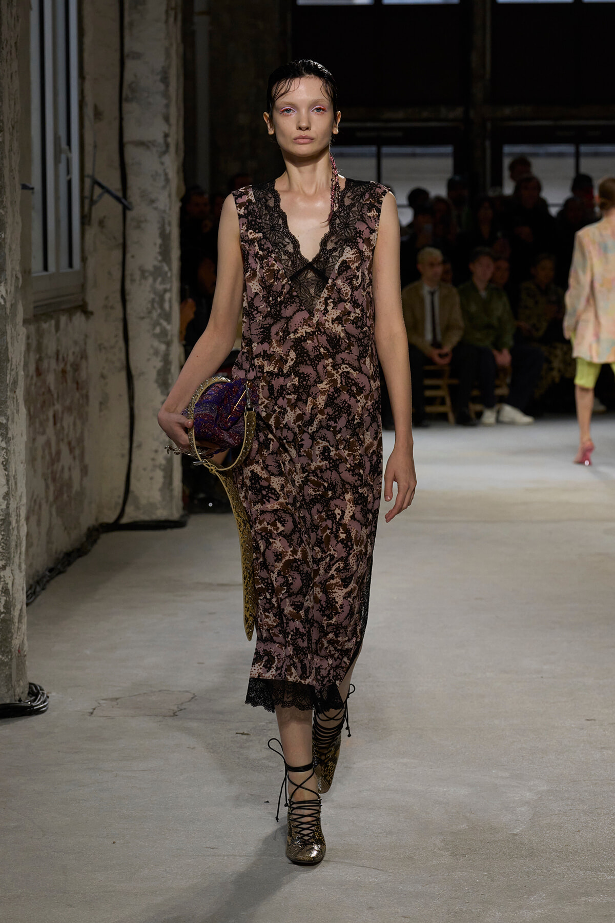 Model walks a fashion runway in a pink-brown lace slip dress, holding a metallic purple bag, in an industrial venue