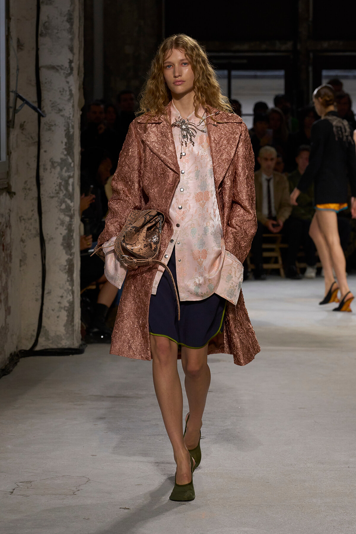 Model walking runway in a pink textured coat over a pale floral blouse and navy shorts, carrying a snakeskin bag and wearing olive-green heels.