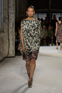 Model walks the runway in a loose, one-shoulder black-and-white animal-print top paired with pink lace-trimmed shorts, finished with metallic lace-up heels, in an industrial venue with an audience in the background.