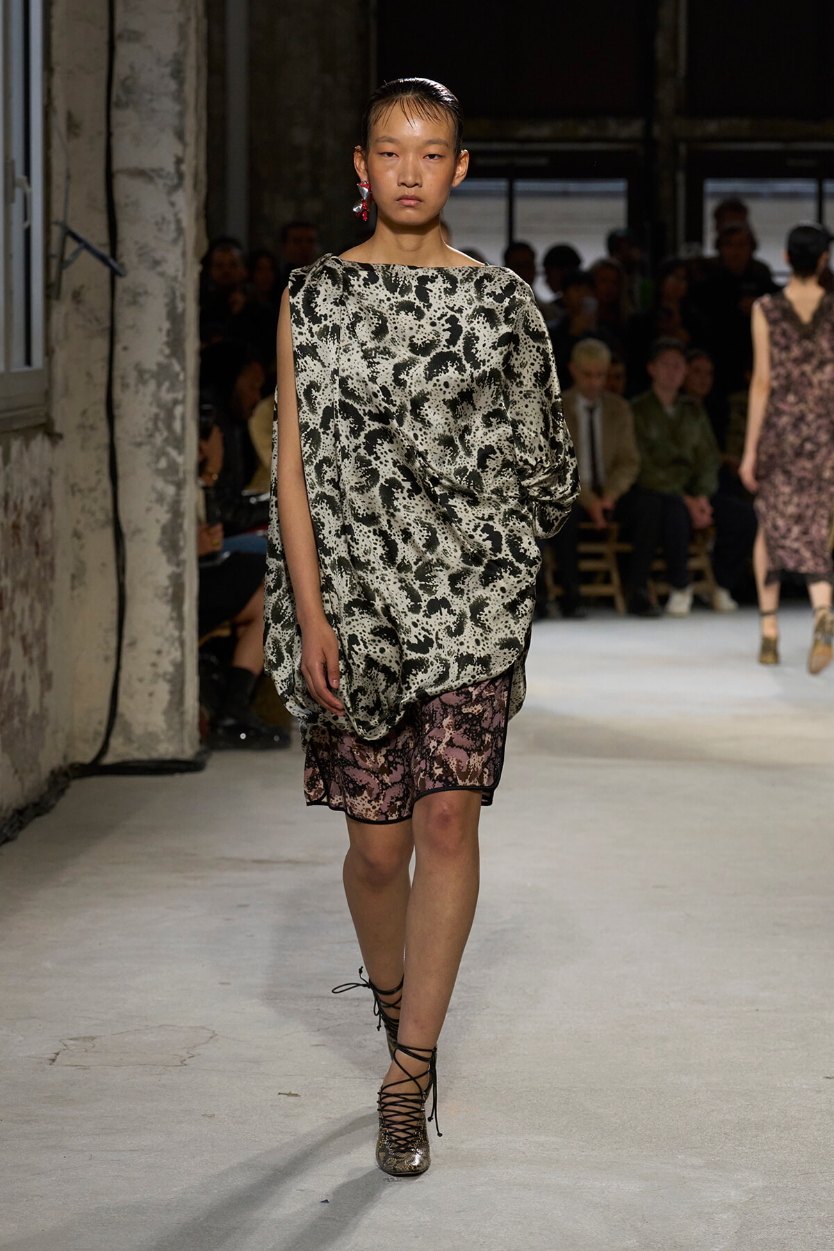 Model walks the runway in a loose, one-shoulder black-and-white animal-print top paired with pink lace-trimmed shorts, finished with metallic lace-up heels, in an industrial venue with an audience in the background.