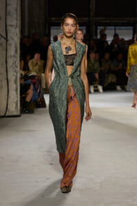 Model walks a fashion runway in a textured green sleeveless coat over a black lace top and orange patterned wide-leg pants, audience in background