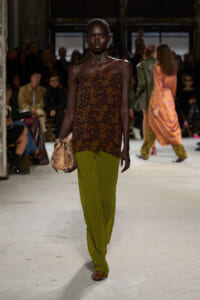 Model walking a fashion runway in a strapless brown lace top and olive green studded pants, carrying a snake-print clutch.