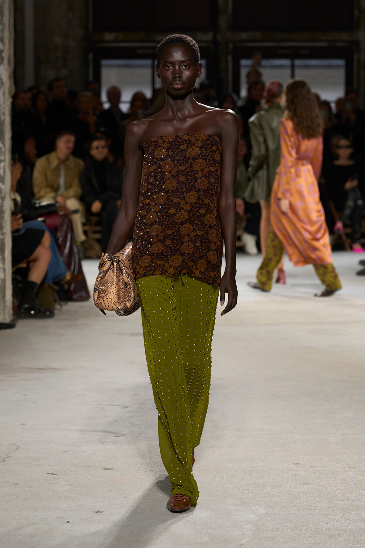 Model walking a fashion runway in a strapless brown lace top and olive green studded pants, carrying a snake-print clutch.