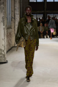 Model walking runway in olive green leather jacket with high ruffled collar, burgundy and olive patterned pants, and snakeskin bag; bold pink eye makeup; industrial venue with spectators in background.
