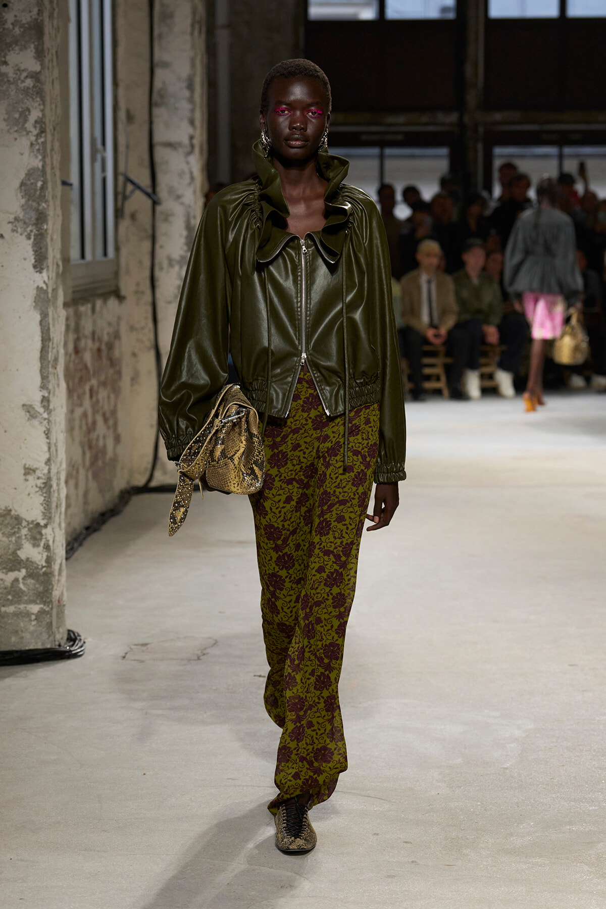 Model walking runway in olive green leather jacket with high ruffled collar, burgundy and olive patterned pants, and snakeskin bag; bold pink eye makeup; industrial venue with spectators in background.