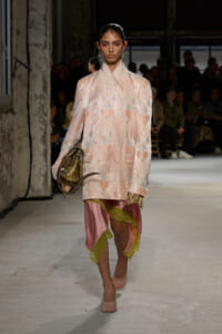 Model on a runway wearing a pale pink floral coat with a high collar, carrying a snakeskin handbag over a pink satin dress with lime lace trim.
