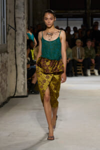 Model walks the runway in a teal beaded camisole paired with a gold brocade wrap skirt featuring a front slit; industrial-looking venue with seated audience in the background.