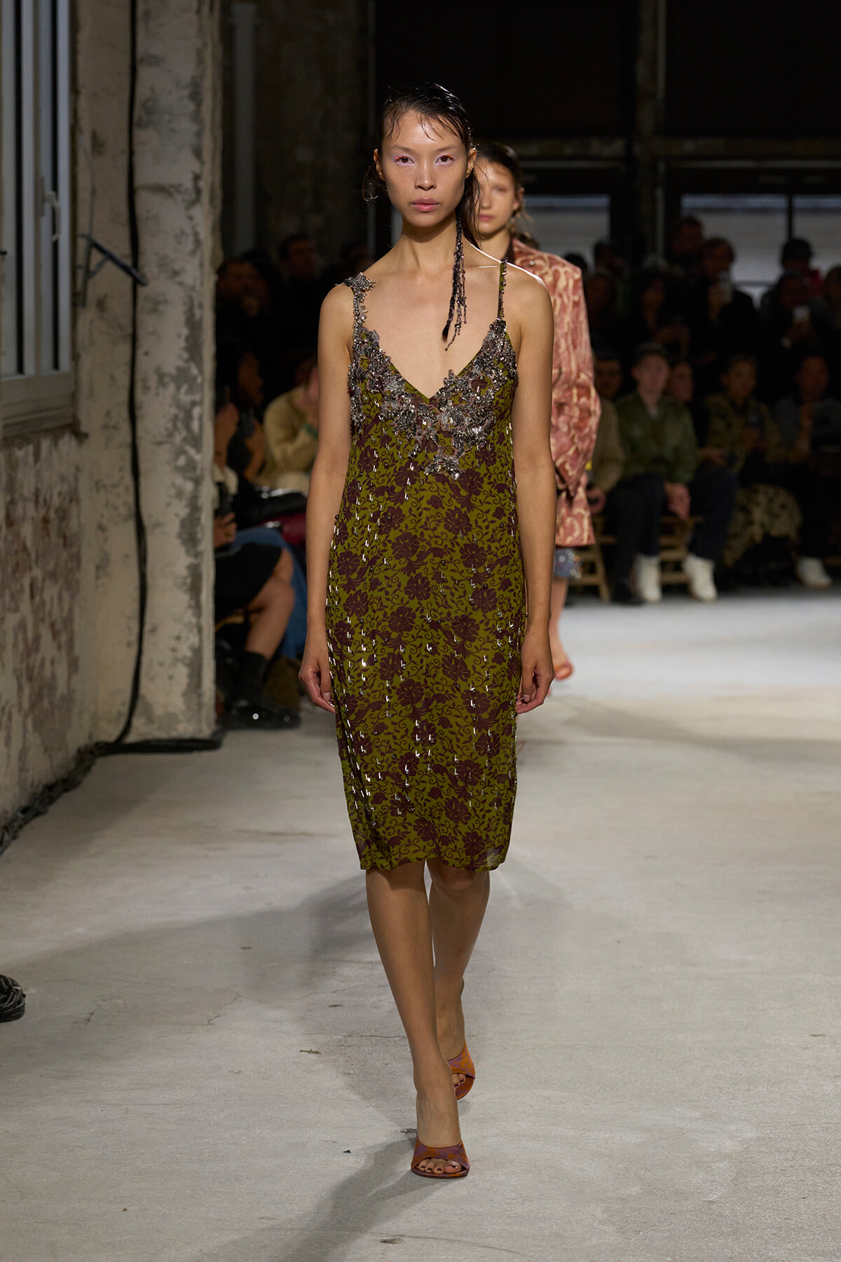 Model walks the runway in a knee-length olive green dress with silver sequin floral embroidery and thin straps, backstage audience visible behind.