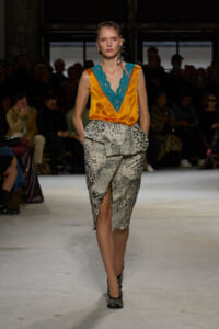 Runway model in an orange satin wrap top with turquoise lace V-neck and a snakeskin-print skirt, walking toward the camera with audience in the background.
