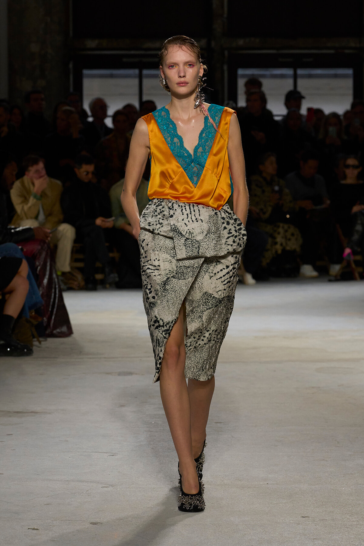 Runway model in an orange satin wrap top with turquoise lace V-neck and a snakeskin-print skirt, walking toward the camera with audience in the background.