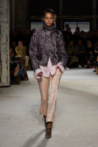 Runway model wearing a purple animal-print jacket over a pink shirt and pastel floral pants, with audience in the background.