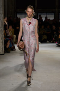Model walking a runway in a pale pink floral jacquard sleeveless dress with front buttons and a thigh-high slit, holding a metallic bronze bag with a curved handle.