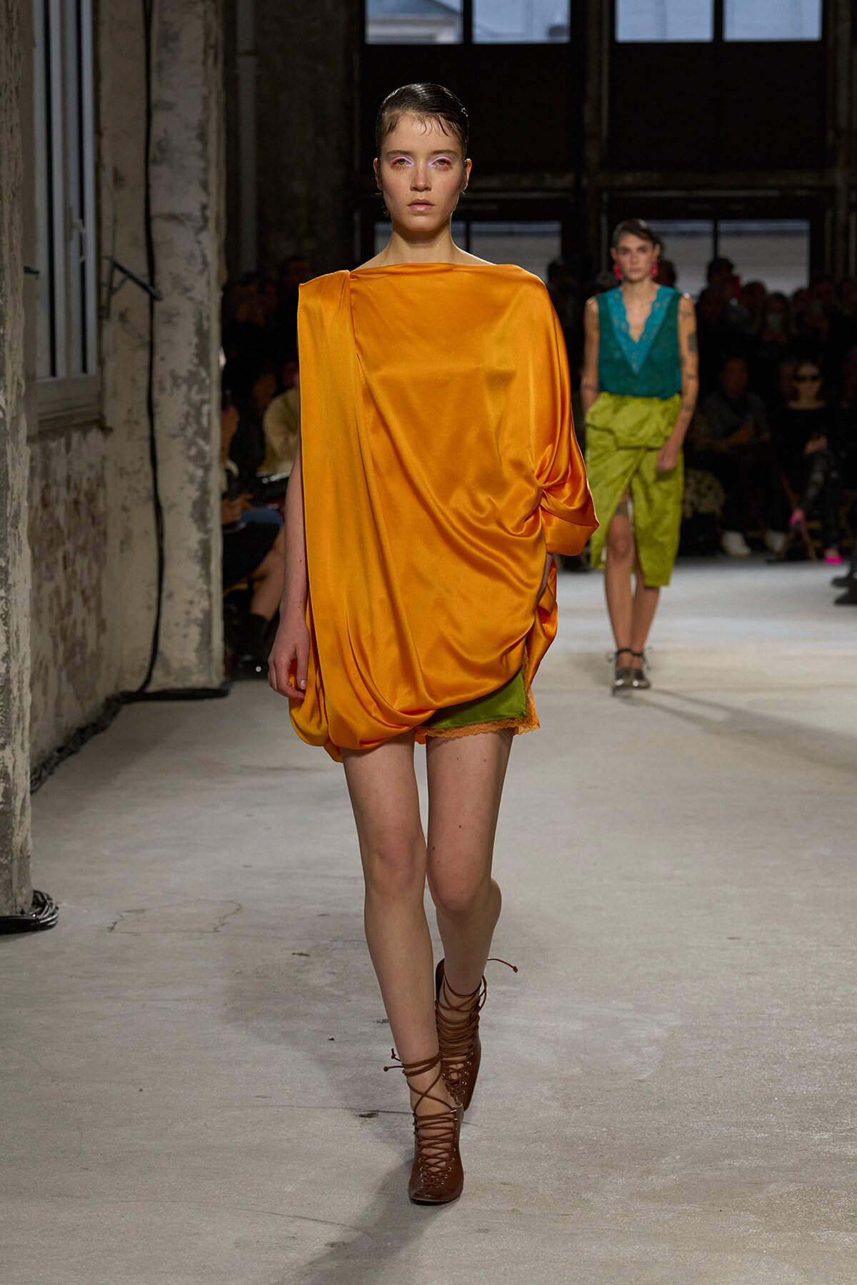 Runway model wearing a bright orange satin draped mini dress with lime-green shorts peeking underneath and brown lace-up boots in a fashion show setting.