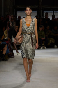 Model walks a runway in a snakeskin-print sleeveless dress with a brown studded belt, pink heels, and a pink clutch.