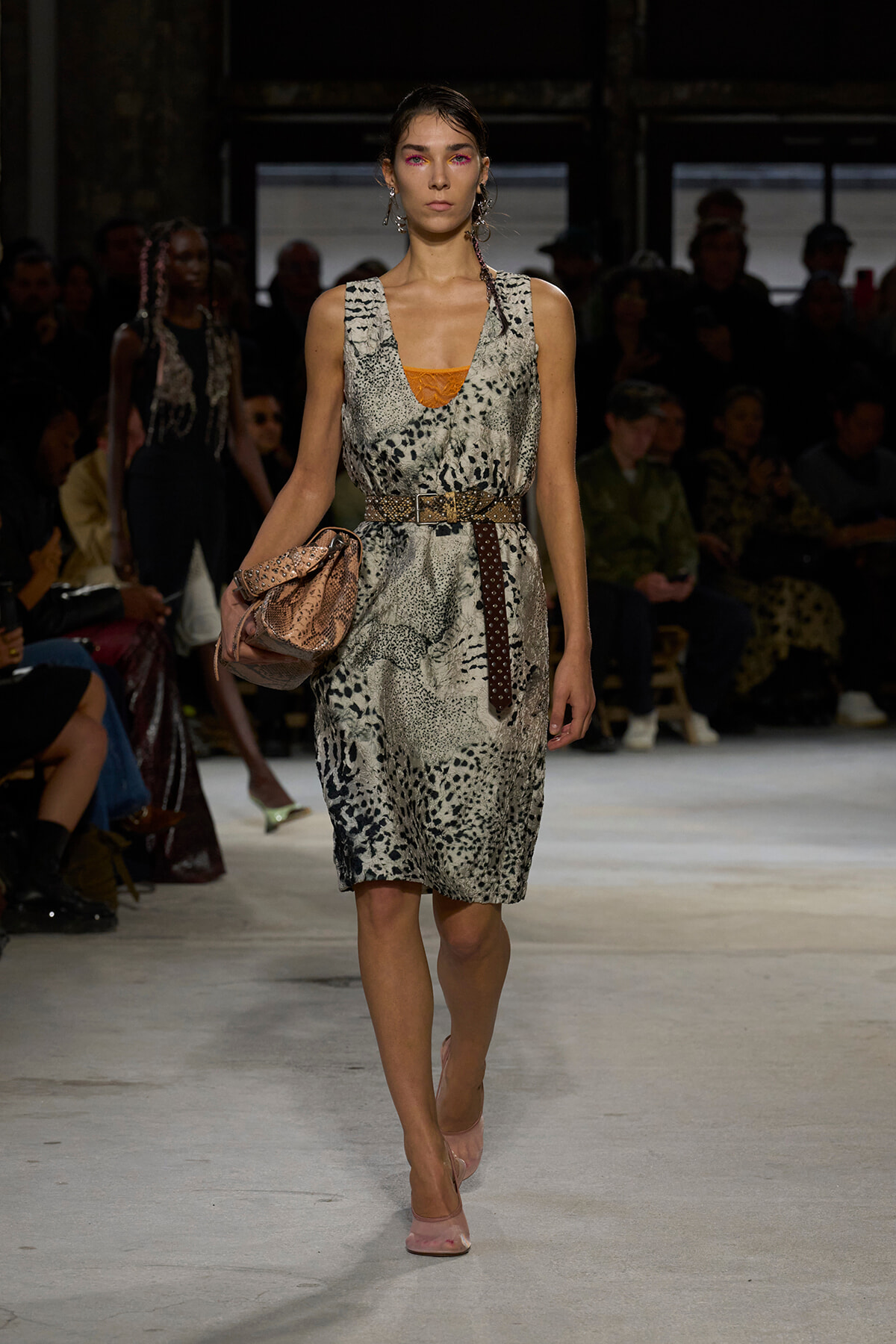 Model walks a runway in a snakeskin-print sleeveless dress with a brown studded belt, pink heels, and a pink clutch.