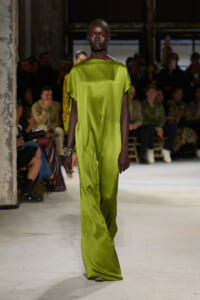 Female model walks a runway in a bright lime‑green satin jumpsuit; audience seated in the background.