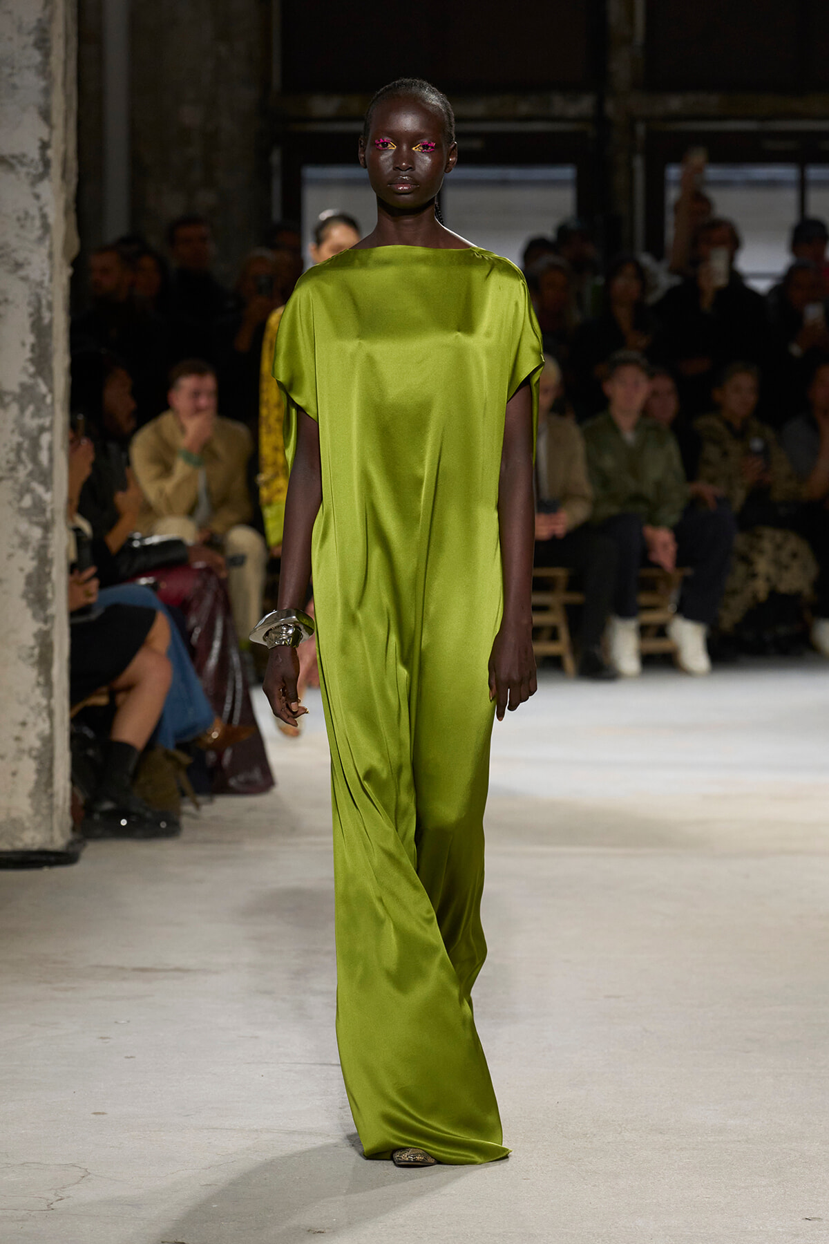 Female model walks a runway in a bright lime‑green satin jumpsuit; audience seated in the background.