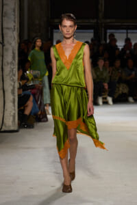 Model walks the runway in an olive green satin dress with orange lace trim and a V-neckline.