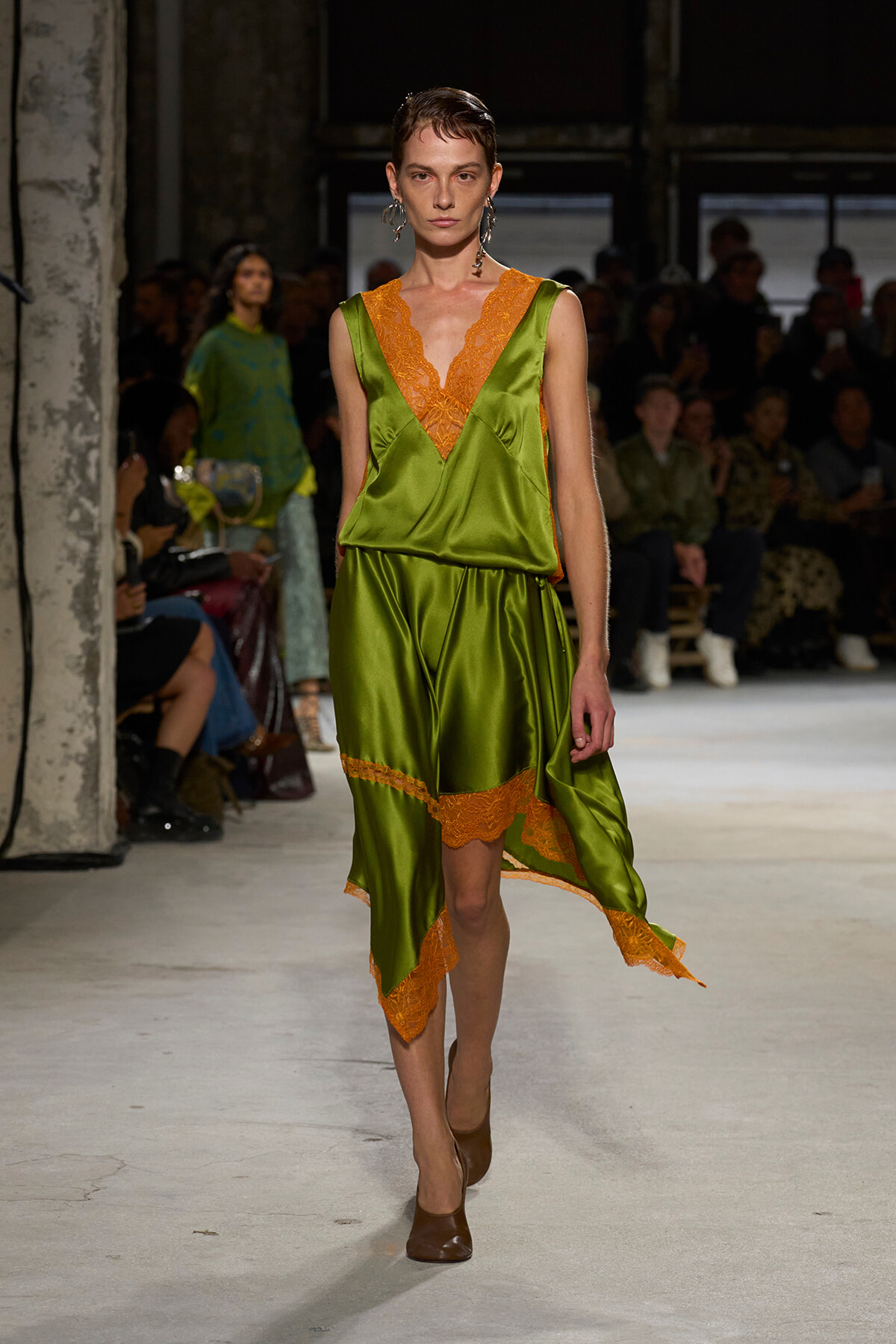 Model walks the runway in an olive green satin dress with orange lace trim and a V-neckline.