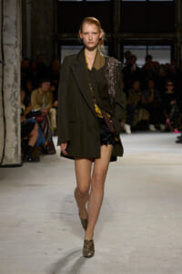 Model walking a runway in an oversized pinstripe blazer, with a brown belt and snake-print ankle boots, on a concrete catwalk with an audience in the background
