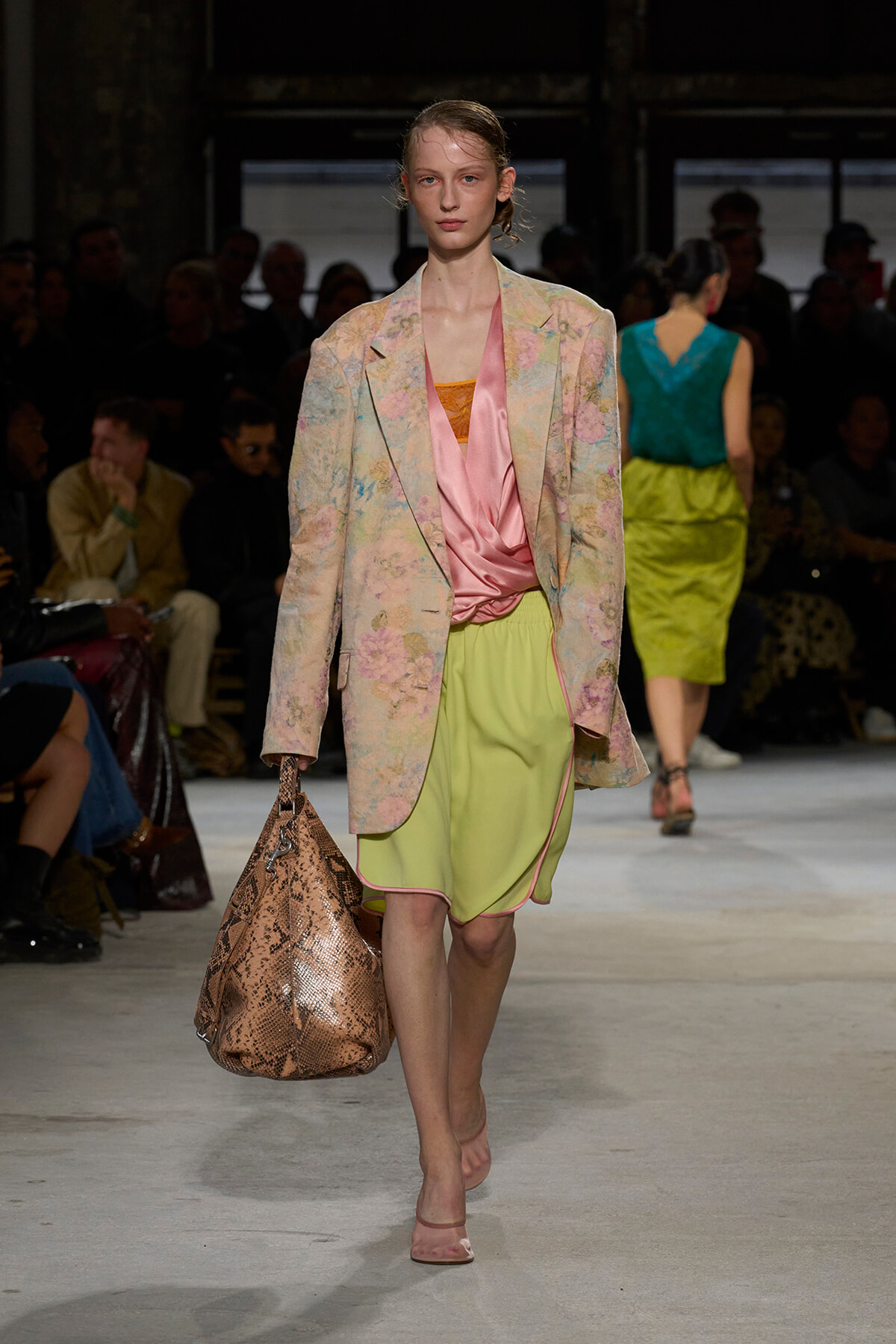 Model walks the runway in a pastel floral blazer, pink satin top, and lime shorts, carrying a snakeskin bag.