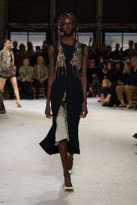 Runway model in a sleeveless black dress with an intricate metallic beaded vest and pale green heels, walking on a fashion show catwalk.