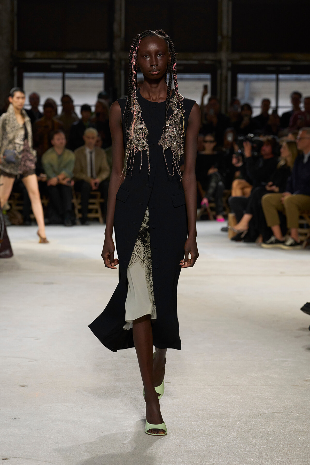 Runway model in a sleeveless black dress with an intricate metallic beaded vest and pale green heels, walking on a fashion show catwalk.