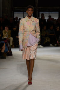 Model walks the runway in a pastel floral suit with a matching lavender blouse and peach shorts, red heels, against a dark audience backdrop.